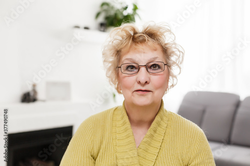 Smiling middle aged mature grey haired woman looking at camera, happy old lady in glasses posing at home indoor, positive single senior retired female sitting on sofa in living room headshot portrait