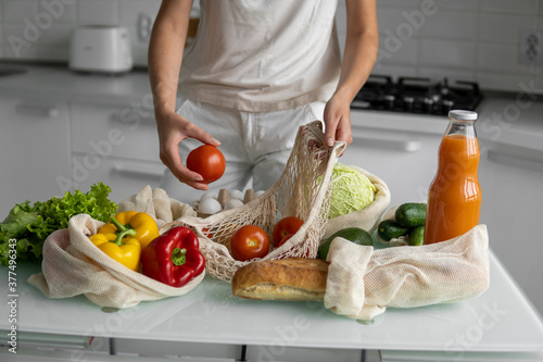 Woman hand holding Reusable eco textile grocery bag with vegetables and takes red tomato out. Zero waste and plastic free concept. Girl is holding mesh cotton shopper with vegetables.