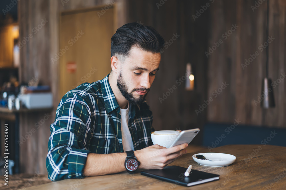 Bearded man drinking coffee in cafe and surfing in internet on smartphone