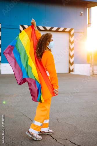 African American lesbian woman wearing a protective mask posing during quarantine of coronavirus pandemic with lgbt pride flag. Covid 2019. LGBT transgender rainbow concept.