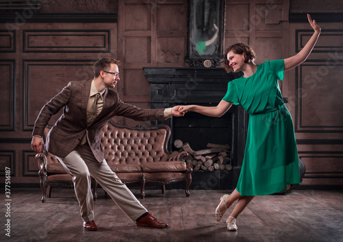 a young couple dancing swing in a retro hall in front of a fireplace and a leathern sofa; the woman wears a green dress