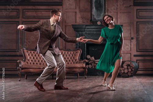 a young couple dancing swing in a retro hall in front of a fireplace and a leathern sofa; the woman wears a green dress, she smiles
