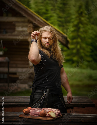 Brutal long hair man prepare meat near forest house