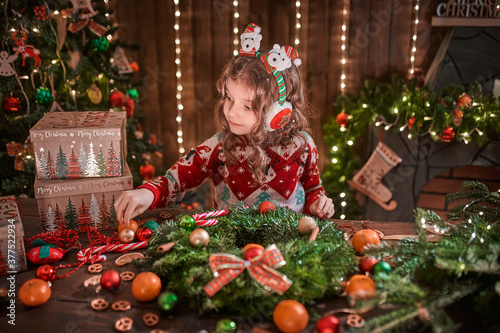 Little girl decorating coniferous wreath near Christmas tree in the decorative interior. Christmas and New Year photo.