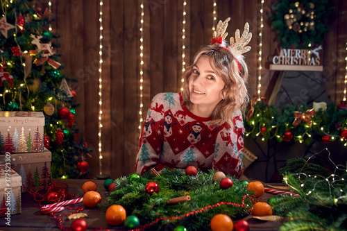 Woman dressed in christmas design sweater, deer horn hoop made handmade christmas wreath on table for holiday, looking at camera smiling. Christmas decoration and composition.