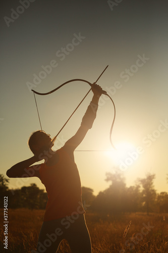 Canvas Print Young Caucasian female archer shooting with a bow in a field at sunset