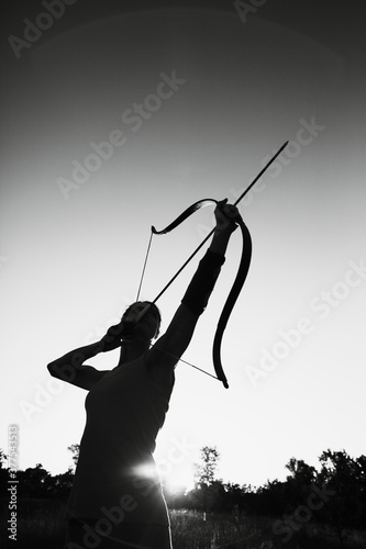 Young Caucasian female archer shooting with a bow in a field at sunset Fototapeta
