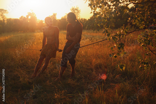 Two men practicing slackline in a field at sunset. Two friends doing sports at sunset.