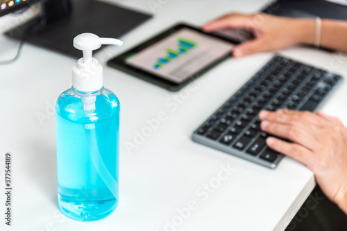 A bottle of alcohol gel on white table work space with background of woman working from home for covit 19 and new normal concept. photo