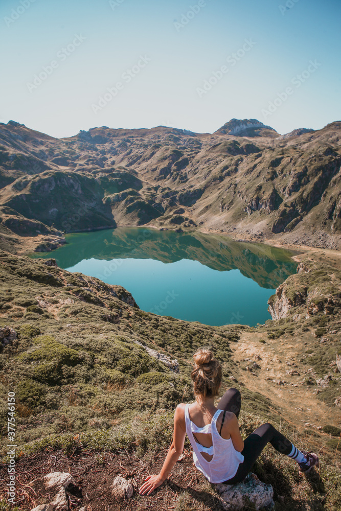 Young woman sitting on the rock watching the beautiful turqouise water of the glacial lake, reflection of the mountain on the surface of the lake in Somiedo natural park in Asturias, Spain