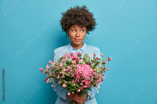 Portrait of attractive curly haired woman holds flowers, dressed in blue shirt and receieves delivery on anniversary. Cute adorable lady in good mood poses for camera with bouquet against blue wall