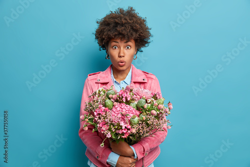 Horizontal shot of good looking woman with curly hair embraces bouquet of flowers, stares impressed and wears pink stylish jacket. Adorable lady enjoys pleasant scent, poses over blue studio wall