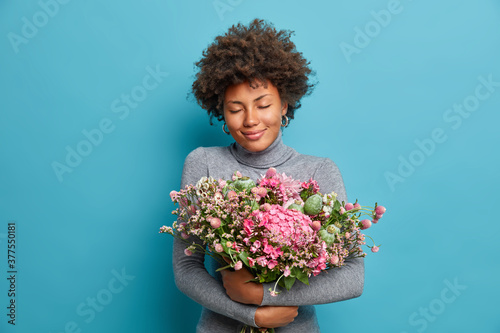 Horizontal shot of pleased dark skinned woman stands with closed eyes and embraces big bouquet, gets proposal from boyfriend. Pretty girlfriend in casual turtleneck enjoys odor of nice flowers