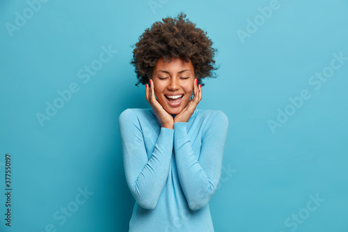 Young happy beautiful African American woman stands joyous with closed eyes, laughs as hears funny joke and touches cheeks. Cheerful positive millenial girl glad to spend free time with friends