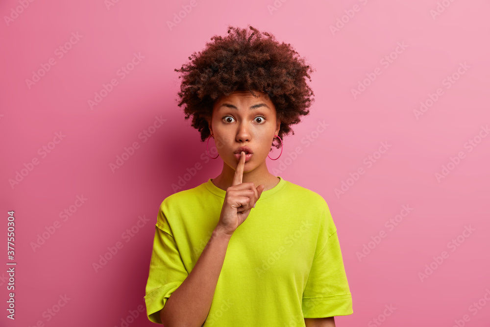 Surprised dark skinned woman makes silence gesture, keeps finger over lips and looks mysterious at camera. Wondered curly millennial girl shows hush sign, poses against rosy background. Body language Surprised dark skinned woman makes silence gesture, keeps finger over lips and looks mysterious at camera. Wondered curly millennial girl shows hush sign, poses against rosy background. Body language
