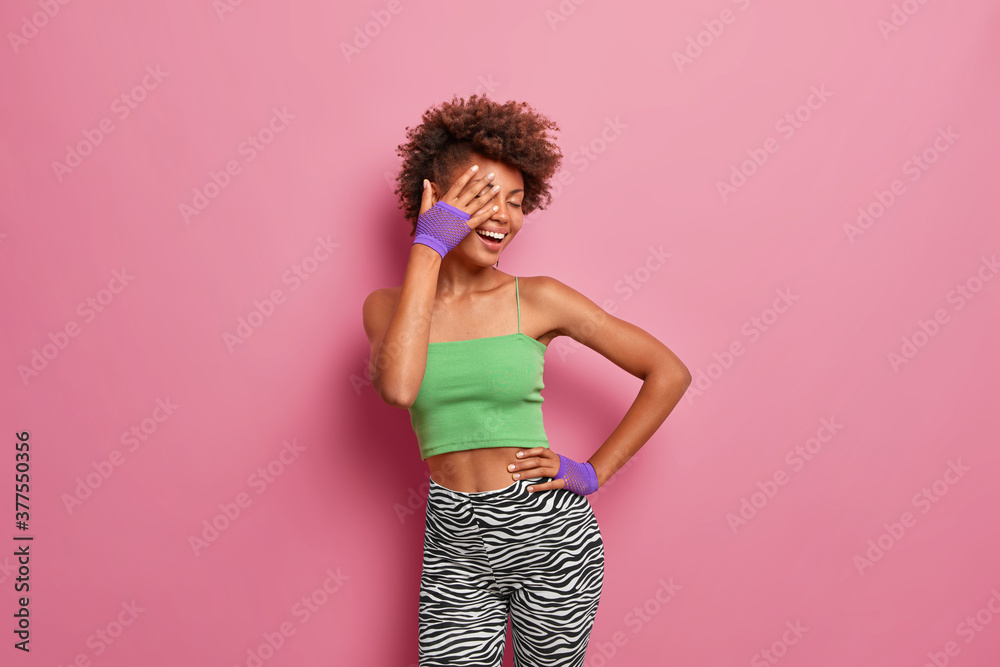 Horizontal shot of optimistic glad curly haired woman makes face palm and laughs positively, dressed in top leggings sport gloves. Sporty ethnic girl has happy mood, poses against rosy background Horizontal shot of optimistic glad curly haired woman makes face palm and laughs positively, dressed in top leggings sport gloves. Sporty ethnic girl has happy mood, poses against rosy background