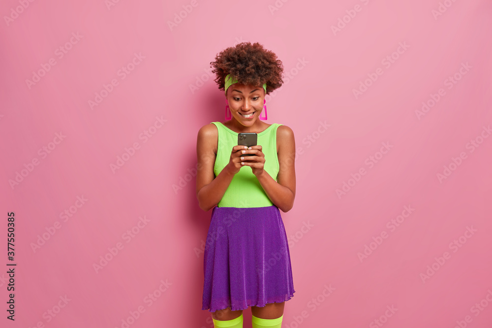 Pleasant looking cheerful female uses mobile phone and free intertnet connection, sends text messages. Glad Afro American woman in trendy clothes poses with modern technology indoor, surfs net Pleasant looking cheerful female uses mobile phone and free intertnet connection, sends text messages. Glad Afro American woman in trendy clothes poses with modern technology indoor, surfs net