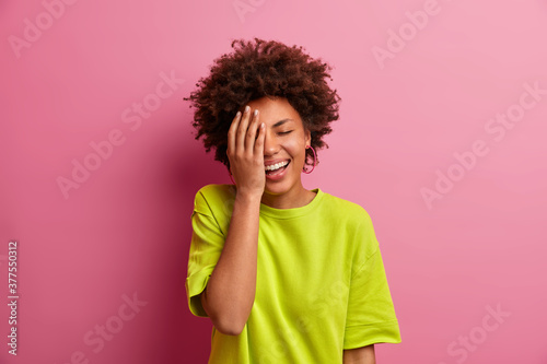 Dark skinned young woman laughs happily and makes face palm  stands with closed eyes. Positive African American girl smiles toothily  wears casual t shirt poses against pink studio background