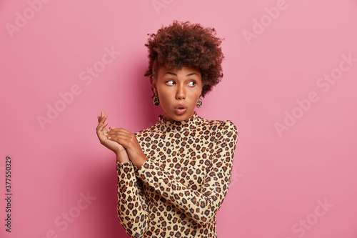 Young stylish pretty woman with Afro hair looks surprisingly aside, keeps mouth opened and rubs hands. Shocked beautiful elegant lady in fashionable leopard outfit poses against pink background