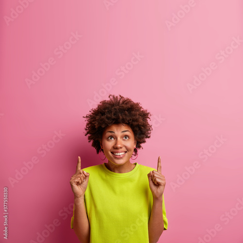 Cheerful smiling beautiful curly haired woman got idea and points above on blank space, advertises something above head. Attractive African American girl in casual wear shows direction upwards