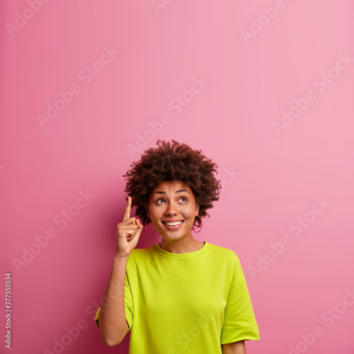 Vertical shot of positive dark skinned woman with curly hair points above on blank space, dressed casually and demonstrates copy space for your advertisement. Pretty girl suggests looking upwards
