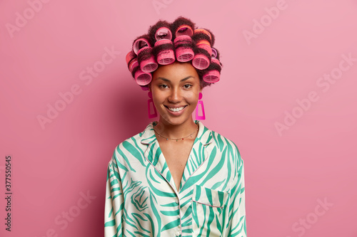 Portrait of pleasant looking young woman with glad expression, wears hair curlers and casual silk dressing gown. Positive lady makes hairstyle on special occasion, poses against pink background