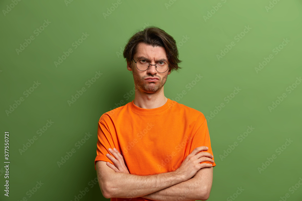 Shot of displeased gloomy man keeps hands crossed over chest and looks from under forehead. Bothered unhappy guy in orange t shirt expresses disagreement or disappointment, poses over green wall Shot of displeased gloomy man keeps hands crossed over chest and looks from under forehead. Bothered unhappy guy in orange t shirt expresses disagreement or disappointment, poses over green wall