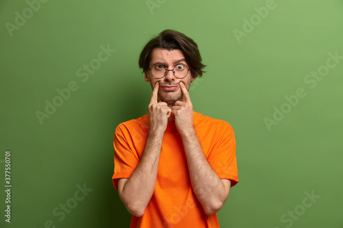 Displeased adult man with trendy hairstyle forces smile and holds fingers near edges of lips, weas casual clothes. Discontent handsome man pretends smiling, stands indoor against green vivid wall