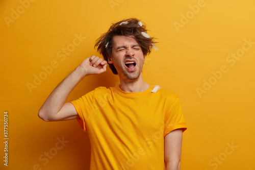 Exhausted lazy man yawns as awakes early in morning has messy hairstyle and wears casual clothes. Fatigue European guy with feathers in hair, wants to sleep and stands against yellow background photo