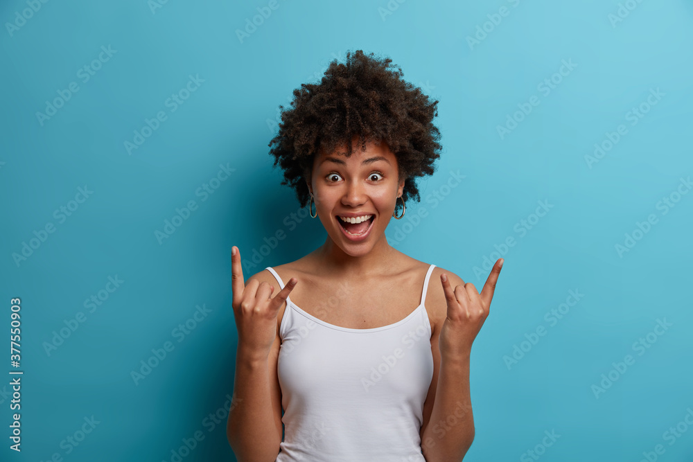 Cheerful emotional makes rock n roll gesture and yells loudly, enjoys concert of favorite band. Joyful ethnic girl shows horns hand sign, poses indoor against blue background. Body language concept Cheerful emotional makes rock n roll gesture and yells loudly, enjoys concert of favorite band. Joyful ethnic girl shows horns hand sign, poses indoor against blue background. Body language concept