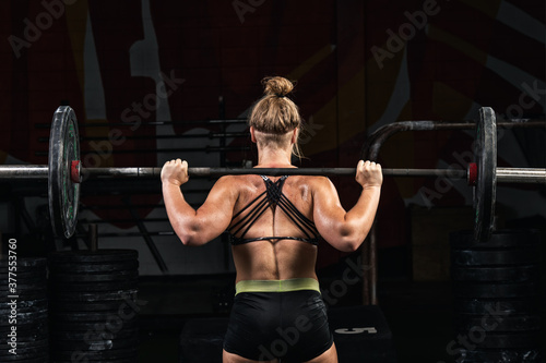 Back view young adult girl doing barbell squats in gym. Woman with muscular body doing lifting exercise. Fit young female lifting barbells looking focused, working out in a gym fitness.