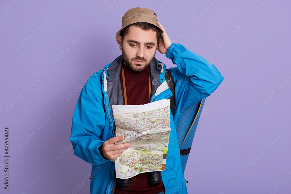 Worried man lost hiking, looking at map with confused facial expression, wearing jacket and hat, keeping hand on head, posing against lilac background. Worried man lost hiking, looking at map with confused facial expression, wearing jacket and hat, keeping hand on head, posing against lilac background.