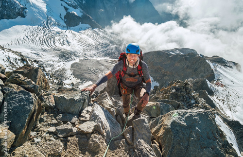 Climber in a safety harness, helmet with backpack asceding a rock wall with Bionnassay Glacier on background and looking at summit during Mont Blanc ascending, France route. Active climbing concept.