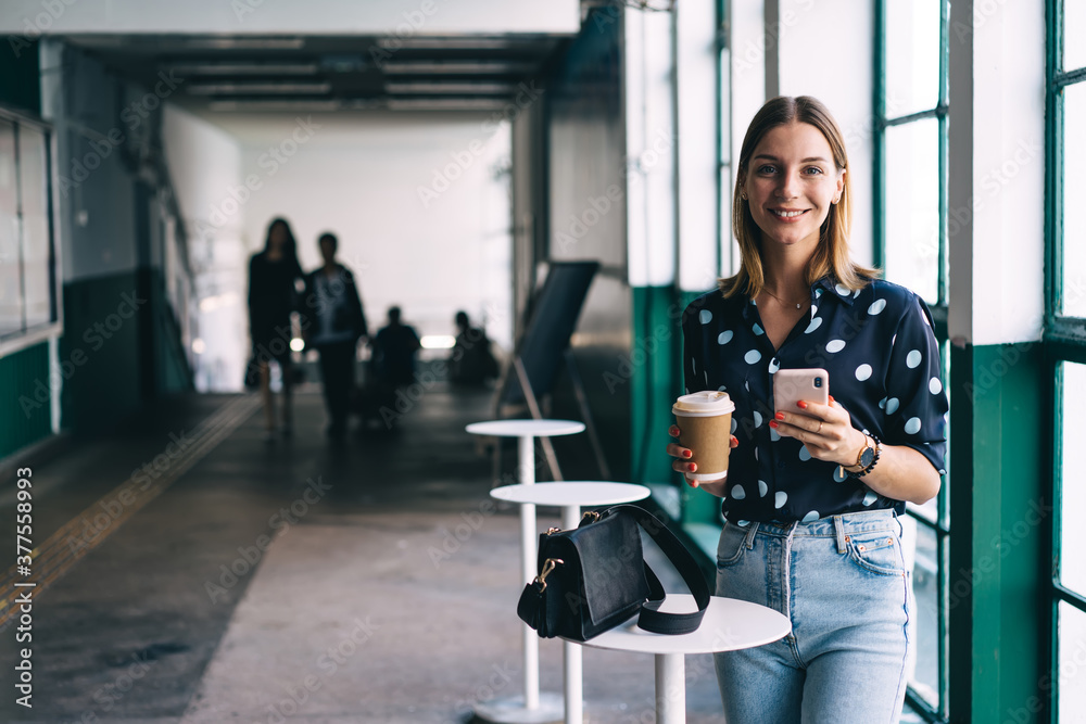 Cheerful female with coffee and smartphone smiling at camera
