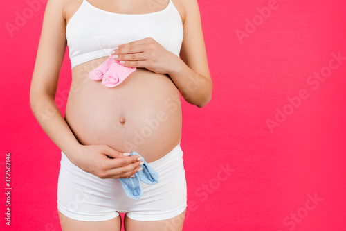Cropped portrait of pregnant woman in white underwear holding baby pink and blue socks against her belly at pink background. Waiting for twins. Is it a boy or a girl Childbirth expecting. Copy space © sosiukin