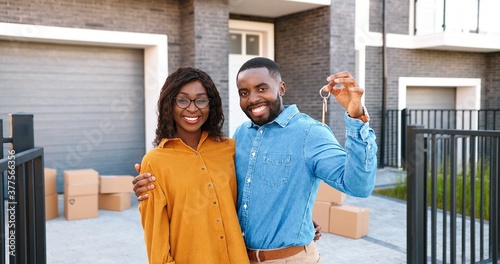 Portrait of happy young African American man and woman smiling to camera and demonstrating key of new home. Outside. Buying real-estate. Moving in concept. Blurred. Cheerful couple settling in.