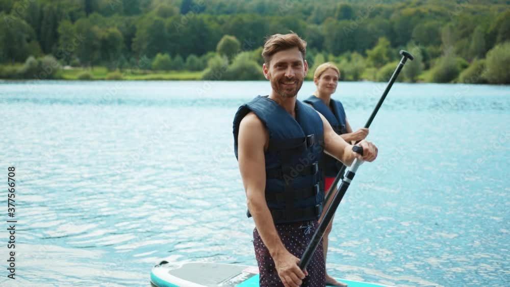 Close up young man look at camera with woman standing on sup board and swims surfing on the lake with a paddle for swimming rowing sport surfer friends slow motion
