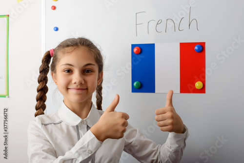 Cheerful young pupil of secondary school in white uniform with pigtails standing in front of white board and showing thumbs up. The flag of France on the blackboard. Concept of learning languages. photo