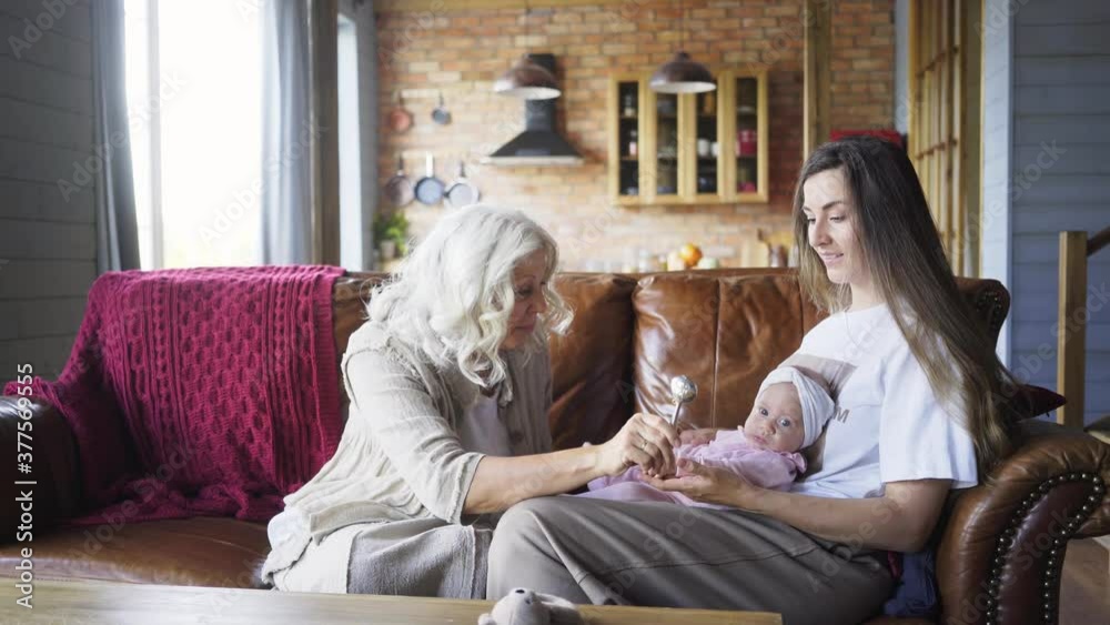 stylish grandmother with long loose grey hair plays with granddaughter in daughter arms sitting on brown leather sofa against stairs at home closeup, side motion shot stylish grandmother with long loose grey hair plays with granddaughter in daughter arms sitting on brown leather sofa against stairs at home closeup, side motion shot