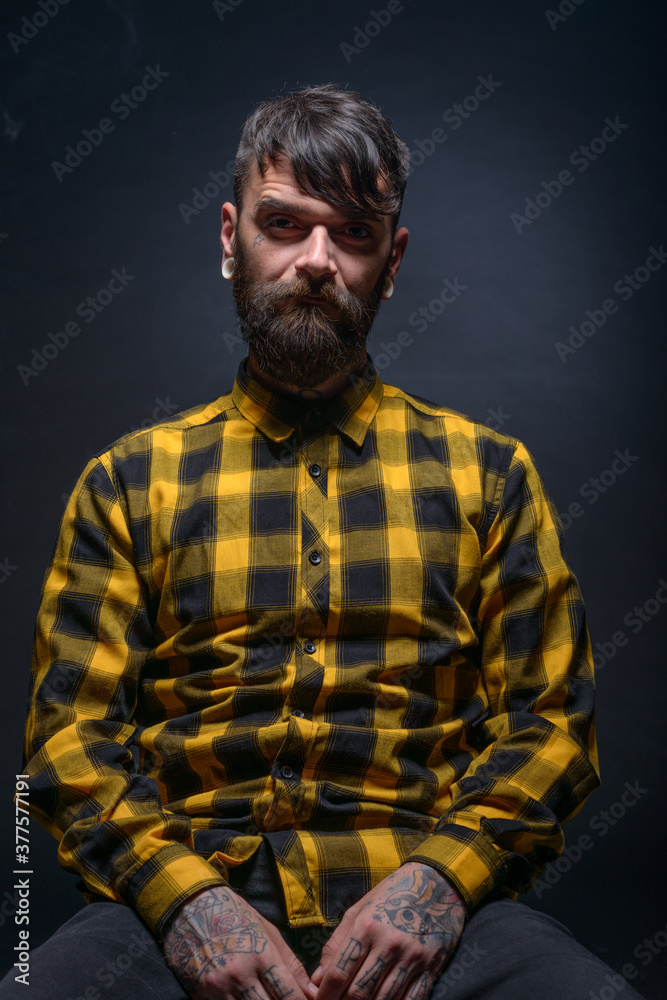 Obraz premium Tattooed man with a beard wearing checkered shirt is posing seated on a chair in a studio