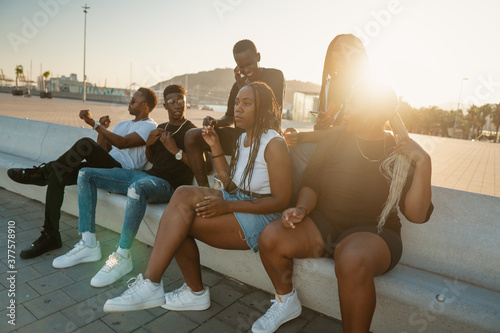 Group of young modern african black friends happily sitting together on the bench of the stairs in bright sunlight enjoying summer vacation © Igor Kardasov
