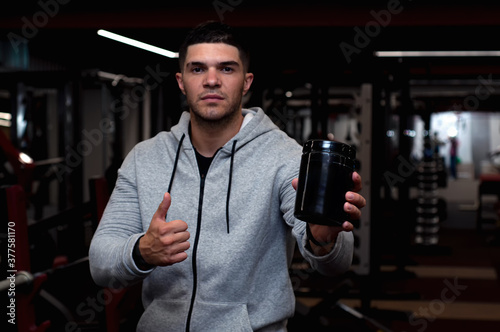 Young athletic guy holding a jar of sports nutrition, gym background