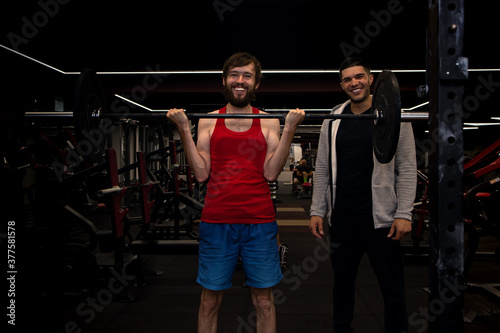 Young man doing exercises with a personal trainer as he lifts a barbell weight to chest height in gym