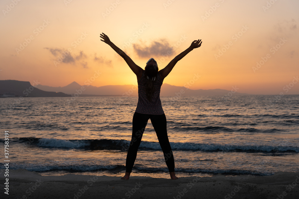 Silhouette, young woman with protective surgical face mask performs yoga stretching exercises at the beach at dusk during covid 19 coronavirus pandemic