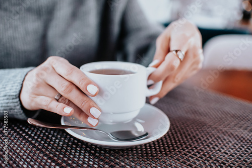 women s hands in a gray sweater with a white manicure hold a white mug of tea at a wooden table