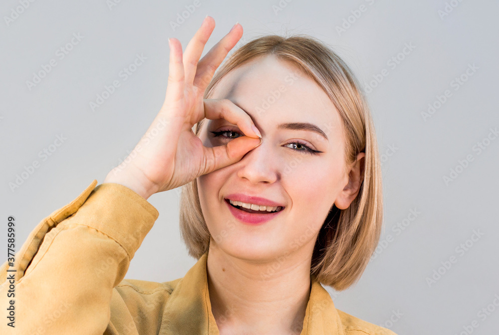 Young woman watching through fingers okay glasses over a grey background Young woman watching through fingers okay glasses over a grey background