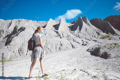 Young blonde woman walking against the white mountains wearing face protective mask for Covid 19 prevention. Young student traveler tourist with backpack. Corona virus and domestic tourism concept