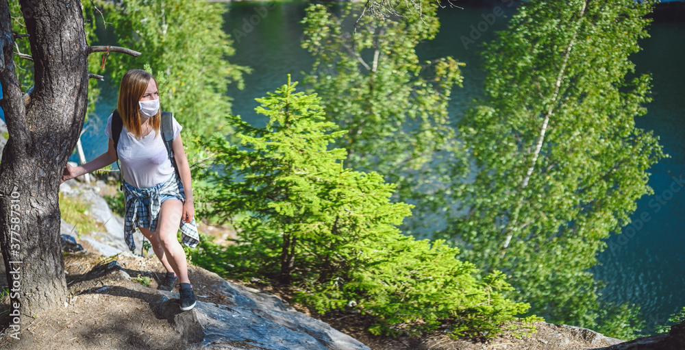Young woman walking against the cliff with green trees and blue mountain lake wearing face protective mask for Covid 19 prevention. Traveler tourist with backpack. Corona virus and domestic tourism. Young woman walking against the cliff with green trees and blue mountain lake wearing face protective mask for Covid 19 prevention. Traveler tourist with backpack. Corona virus and domestic tourism.