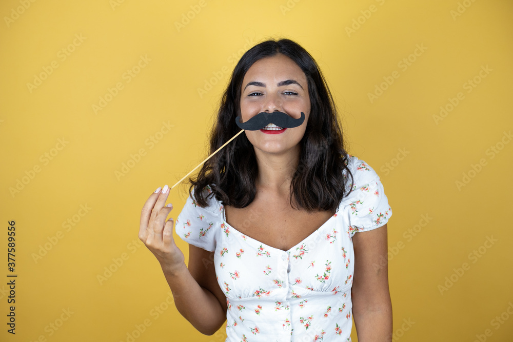 Beautiful woman over yellow background happy with mustache mask Beautiful woman over yellow background happy with mustache mask