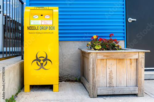 A public drop box for used needles, on a sidewalk by a building. The box is bright yellow, and bears the standard bio-hazard symbol.